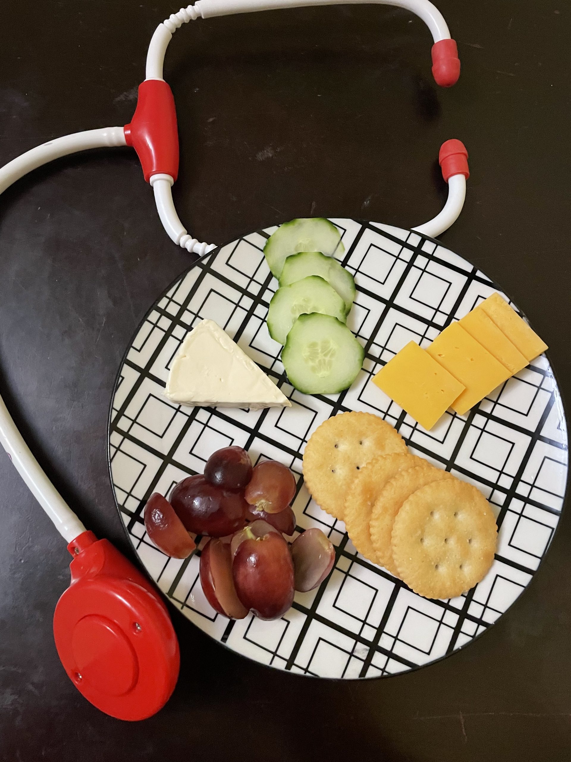 Toddler Snack Plates with sliced cucumbers, cheddar cheese squares, Ritz crackers, cute red grapes, and Laughing Cow cheese wedge arranged with a toy stethoscope, sitting on a brown table.