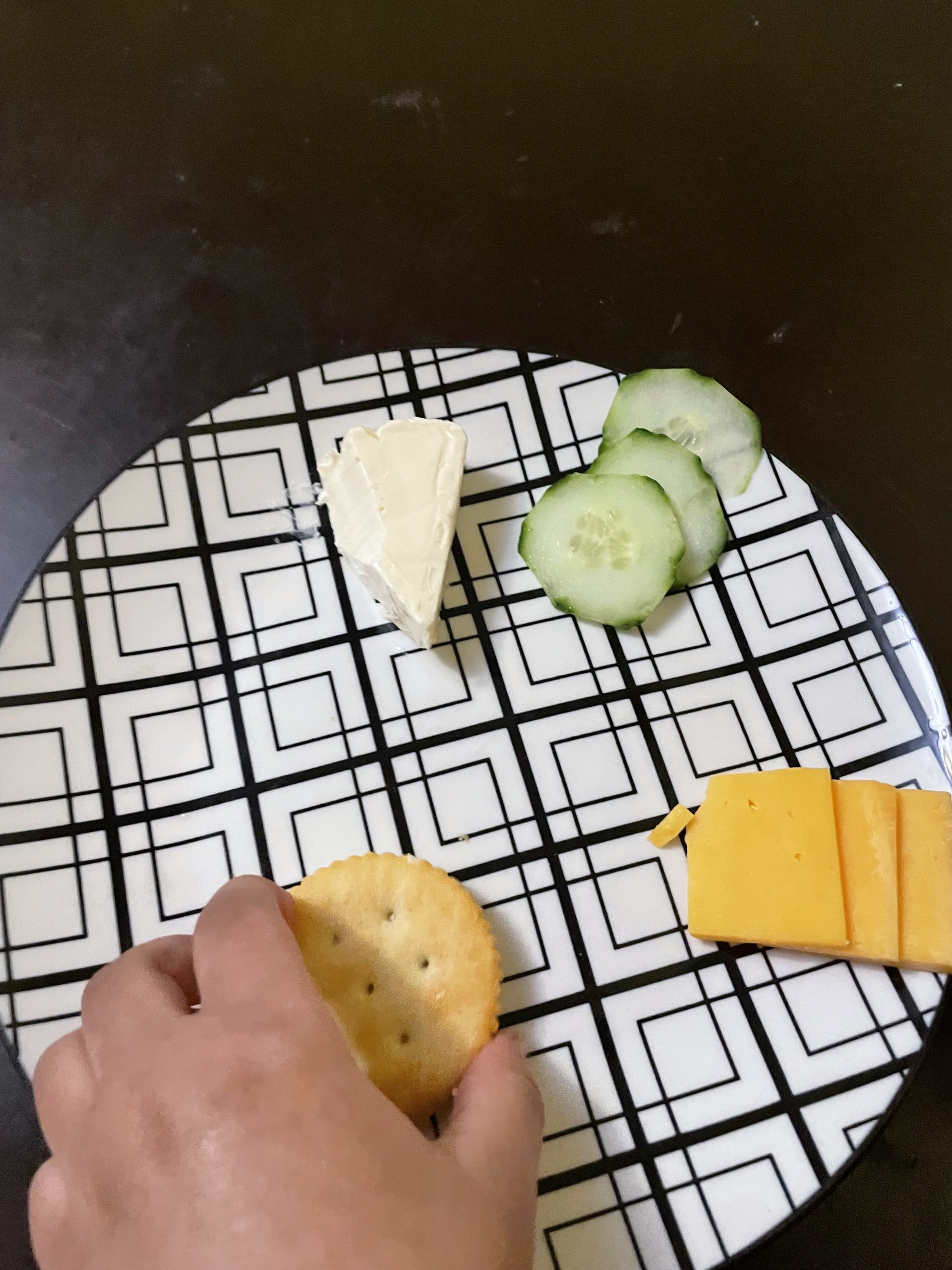 Toddler snack plate with Laughing Cow cheese, cucumber slices, cheddar cheese squares, and a toddler hand picking up a cracker on a dark brown table.