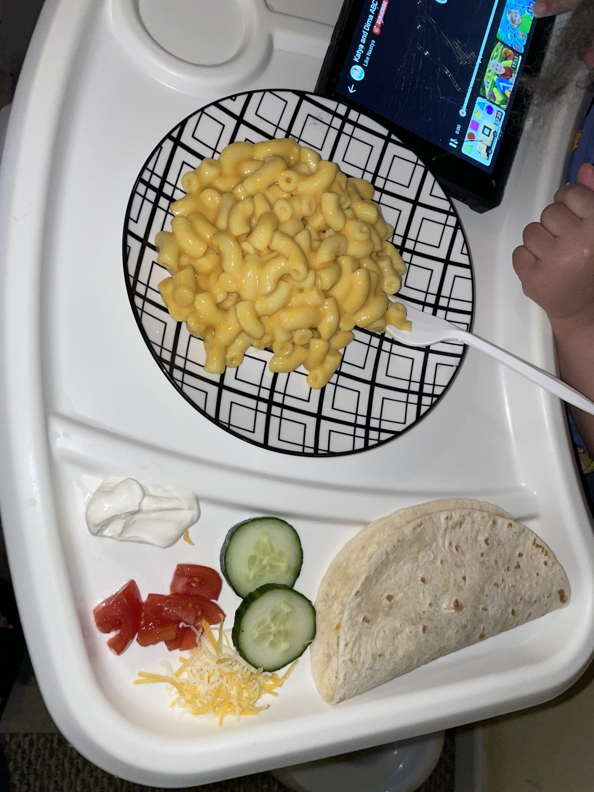 Toddler sitting in highchair during family mealtime with mac and cheese, a taco, cucumber slices, shredded cheese, diced tomatoes, and sour cream on the tray.