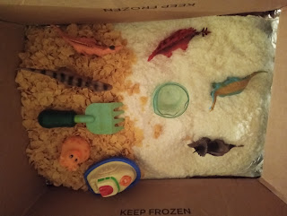 Toddlers' sensory bin with dried potato flakes, cornflakes, toy dinosaurs, a toy boat, a small plastic cup, and a toy rake for hands-on food exploration.