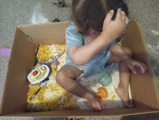 Three-year-old boy sitting in a sensory bin filled with potato flakes, cornflakes, and toys, exploring textures with his hands.