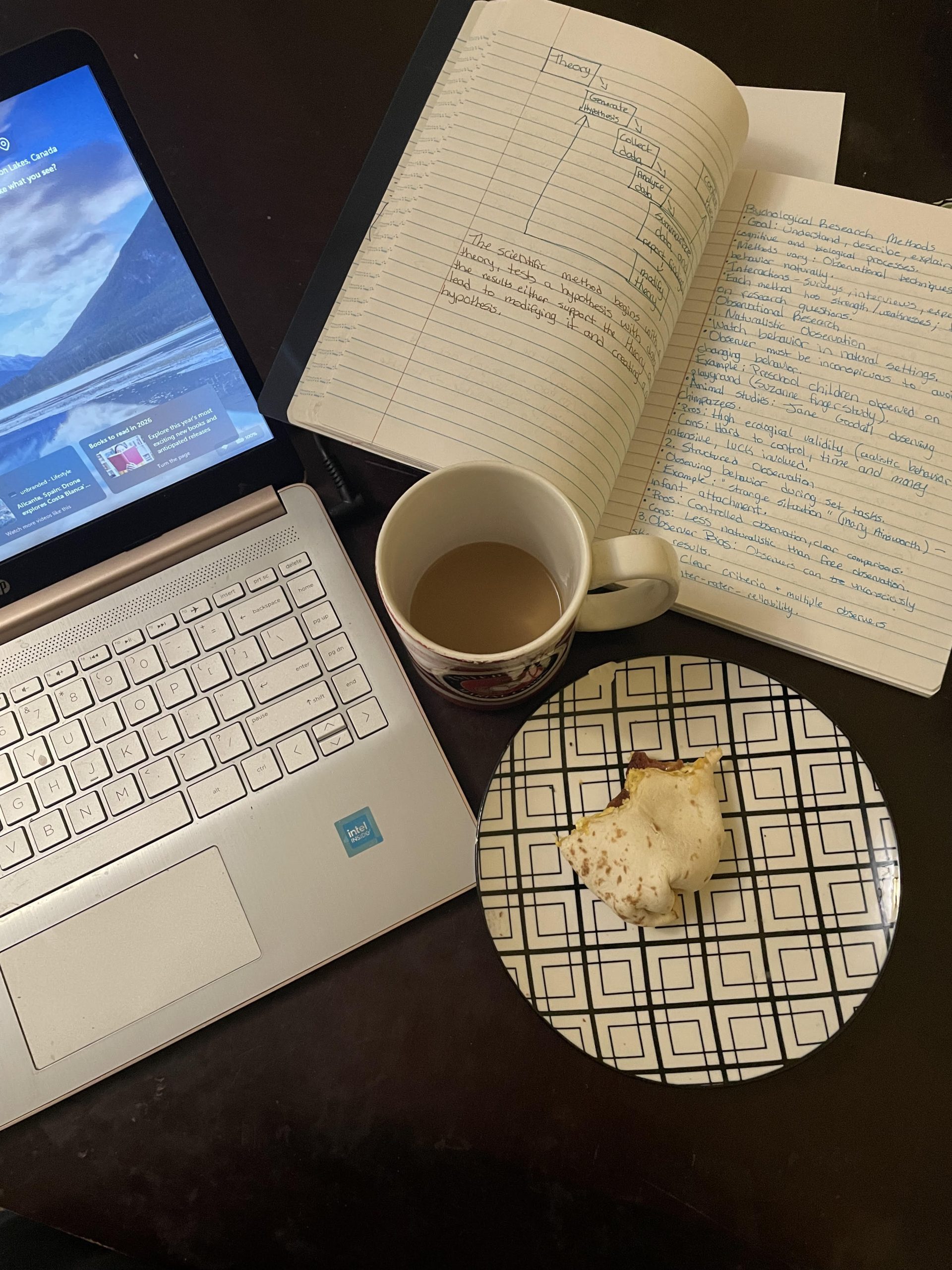 Realistic mom work space with open notebook, laptop, partially eaten breakfast, and coffee on a dark brown table, showing a day in the life with a toddler.